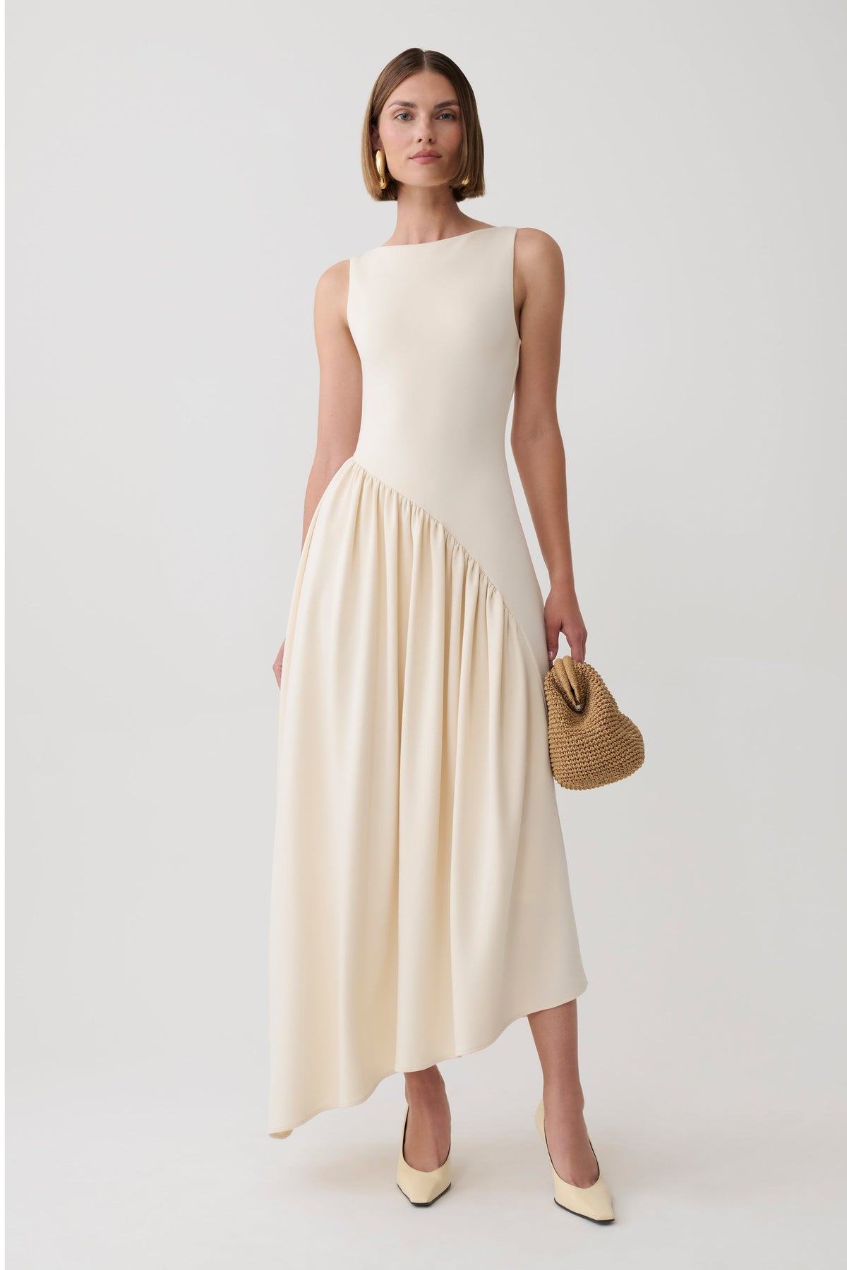 A woman models a sleeveless cream-colored midi dress with a structured bodice and a flowing skirt, holding a small woven straw handbag against a light gray studio background.