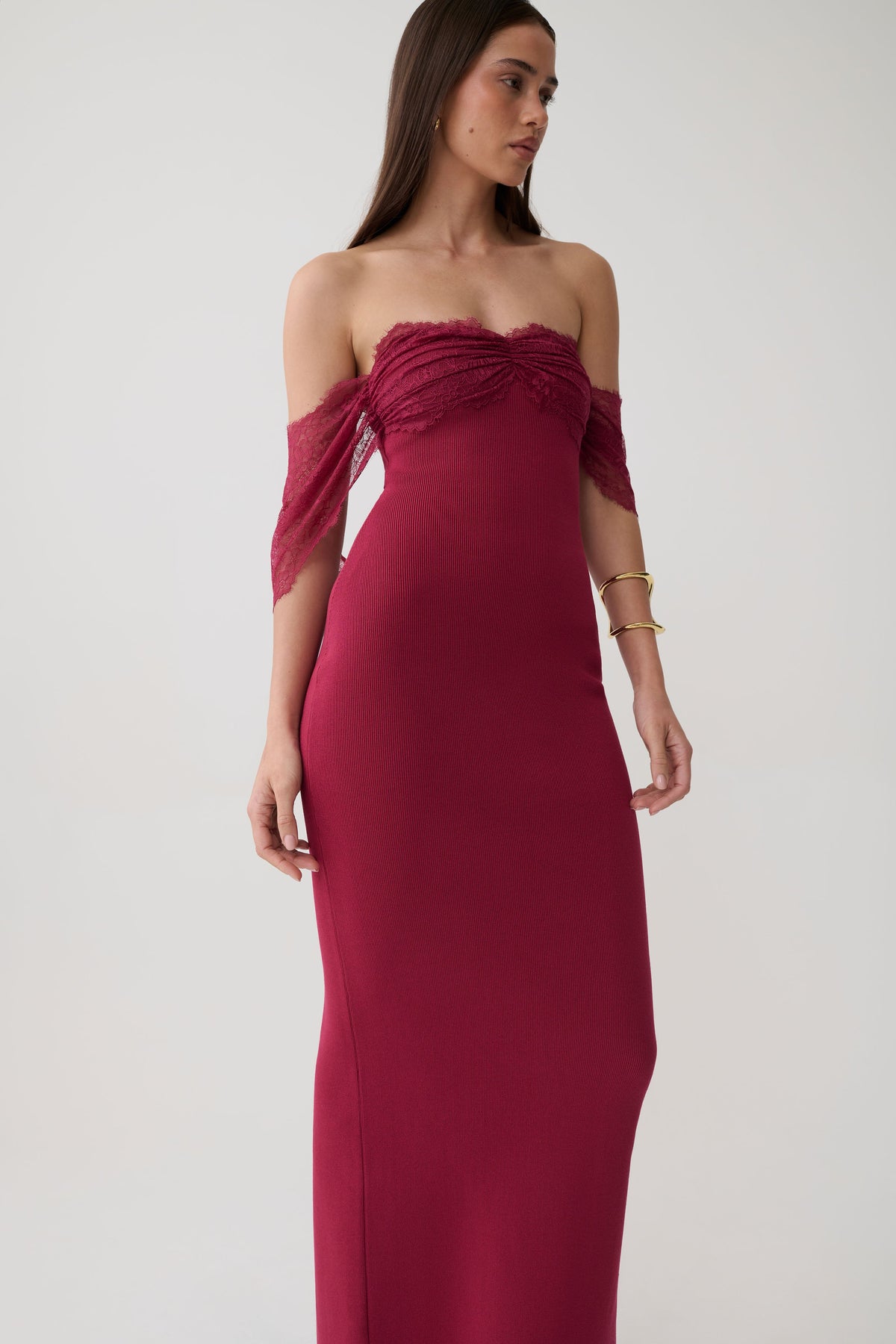 A woman modeling a strapless off-the-shoulder maxi dress in deep burgundy. The dress features a ruched bodice and sheer mesh sleeves, shown against a plain light gray studio background.