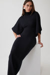 A woman models a black high-neck, long-sleeved knit dress with draped cape-like sleeves, leaning against a white block in a minimalist studio setting.