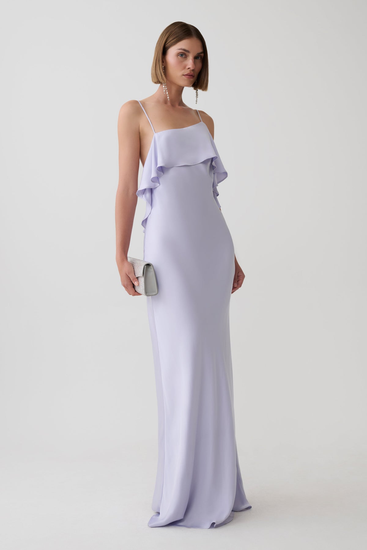 A woman modeling a floor-length lavender slip dress with thin straps and a ruffled neckline, standing against a plain light gray studio background and holding a small light gray clutch.