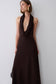 A woman wearing a sleeveless dark brown halter-neck maxi dress with a deep plunging ruffle neckline, standing against a light gray wall.