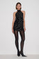 A model stands against a plain light gray studio background wearing a sleeveless black halter mini dress covered in small silver studs, sheer black tights, and black pointed heels.