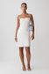 A woman modeling a white sleeveless mini dress with thin straps, paired with a light blue shoulder bag and white strappy heels, against a plain light gray studio background.