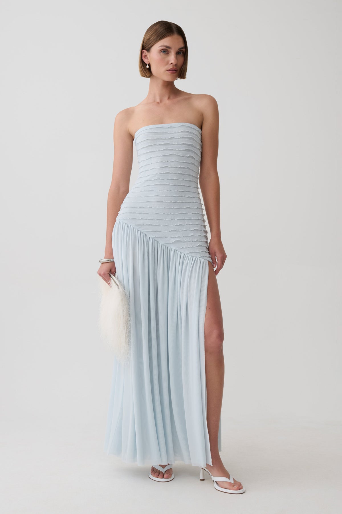 A woman models a strapless light blue pleated maxi dress with a high thigh slit, holding a white feather clutch against a plain light gray studio background.