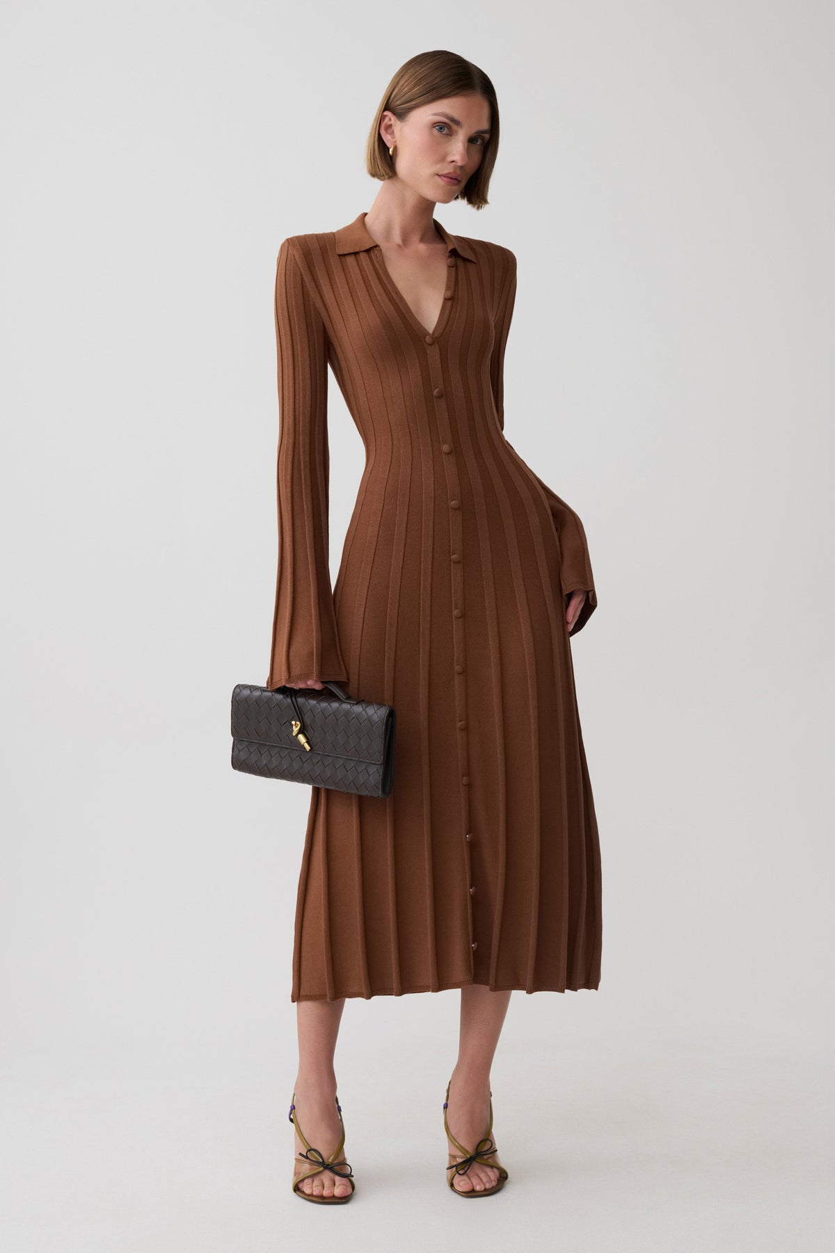 A woman models a brown ribbed long-sleeve midi dress with a button front and V-neckline, holding a black quilted handbag against a light gray studio background.