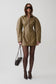 A model wearing a structured, oversized olive-brown mini jacket with a front zipper, styled with black tights, pointed-toe heels, sunglasses, and a small black handbag against a light gray studio background.