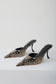A pair of pointed-toe mule heels with a black and beige snakeskin pattern, displayed on a clean white studio background.