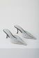 A pair of pointed-toe mule heels in a pale icy blue color, featuring decorative cutout detailing on the sides and slim dark brown stiletto heels, photographed on a clean white studio background.