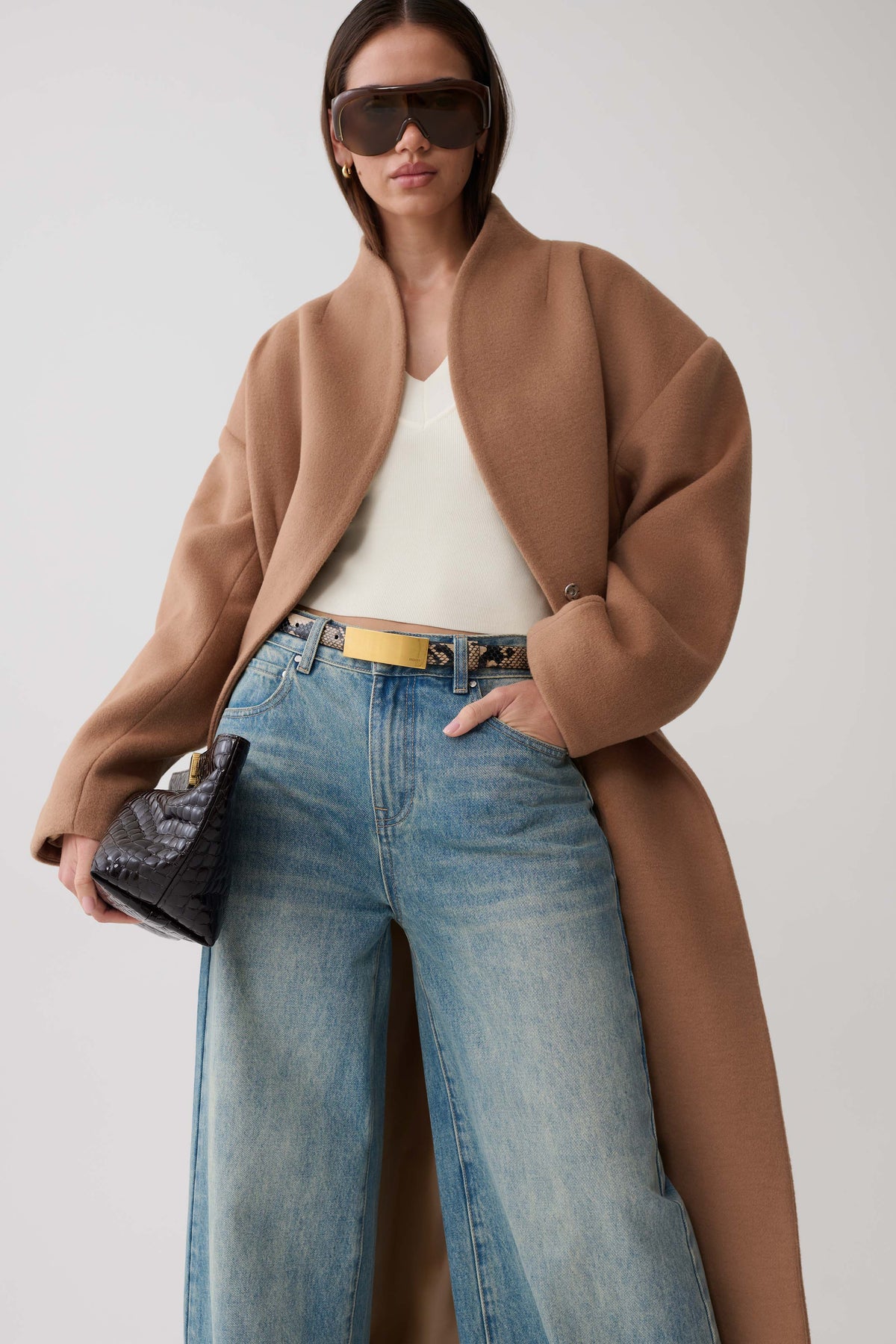 A model wearing a camel-colored wool-blend coat over a white top and wide-leg blue jeans, styled with a patterned belt, sunglasses, and a black clutch bag against a light gray studio background.