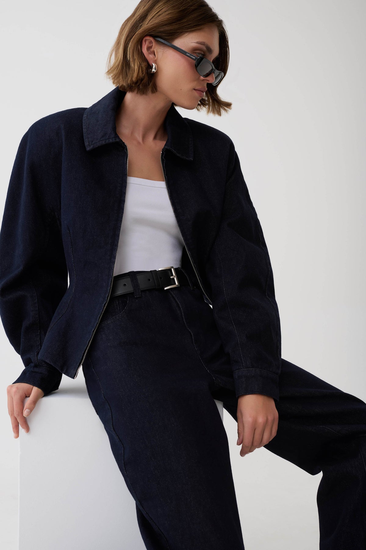A model wearing a dark navy denim jacket over a white tank top and dark navy trousers, accessorized with a black belt and sunglasses, seated against a plain white background.