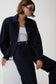 A model wearing a dark navy denim jacket over a white tank top and dark navy trousers, accessorized with a black belt and sunglasses, seated against a plain white background.