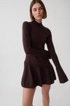 A woman modeling a dark brown ribbed knit mock-neck long-sleeve top with a matching pleated mini skirt, photographed against a light gray studio background.