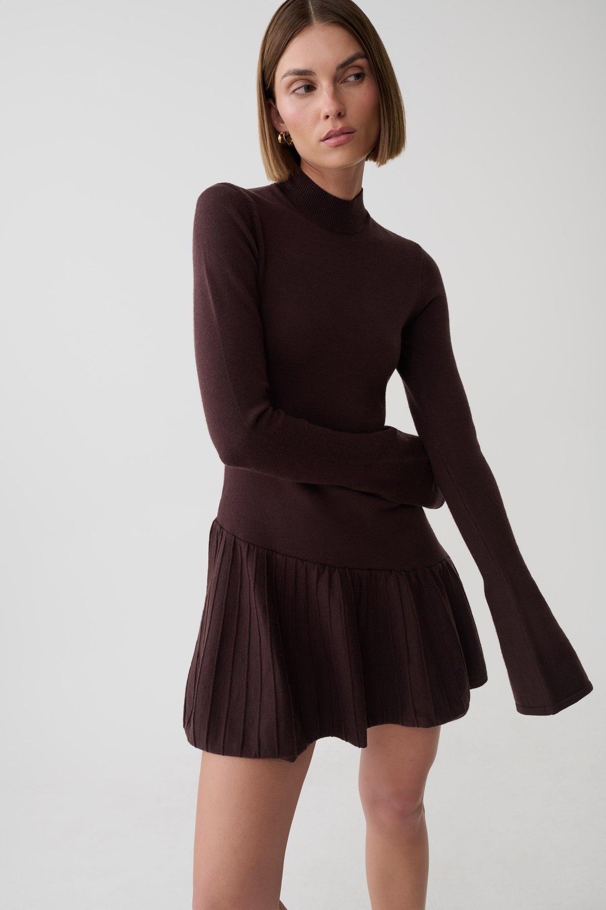 A woman modeling a dark brown ribbed knit mock-neck long-sleeve top with a matching pleated mini skirt, photographed against a light gray studio background.