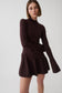 A woman modeling a dark brown ribbed knit mock-neck long-sleeve top with a matching pleated mini skirt, photographed against a light gray studio background.