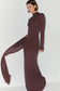 A woman modeling a long-sleeve, high-neck, floor-length dress in a deep plum-brown color, featuring a draped sheer side panel that flows out to the side against a plain white studio background.
