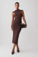 A model wearing a sleeveless, bodycon midi dress in a dark chocolate brown color, styled with a black textured clutch and dark pointed-toe heels, set against a plain light gray studio background.