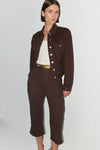 A model wearing a dark brown cropped jacket and matching cropped trousers with gold-tone buttons and a gold belt, styled over a white top against a light gray studio background.