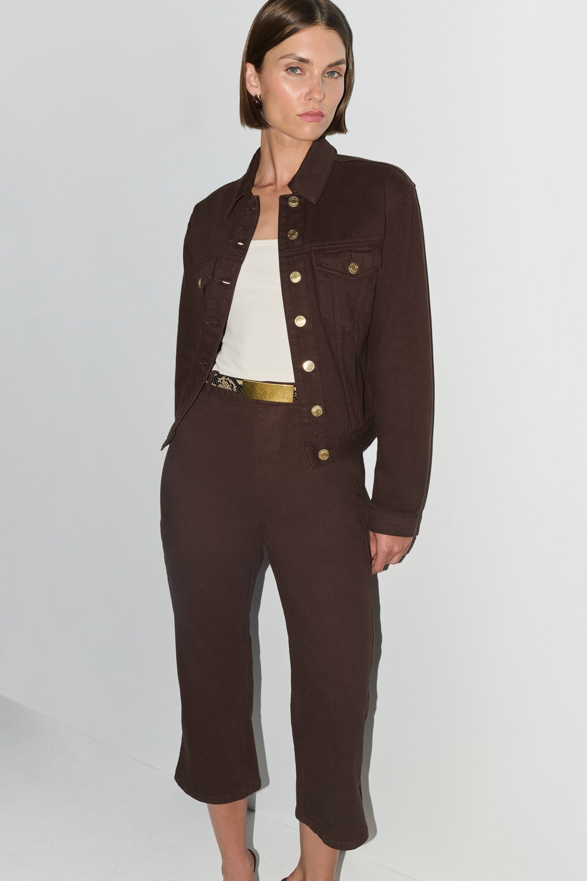 A model wearing a dark brown cropped jacket and matching cropped trousers with gold-tone buttons and a gold belt, styled over a white top against a light gray studio background.