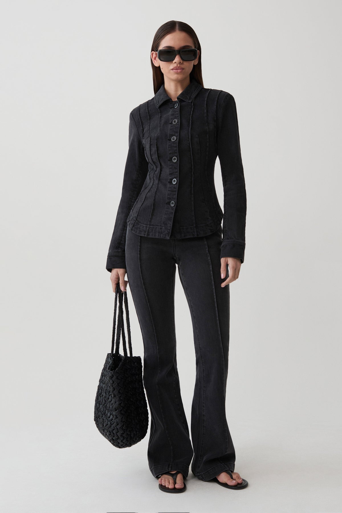 A model stands against a light gray studio background wearing a black textured button-front jacket, black wide-leg pants, black sunglasses, and holding a black woven tote bag.