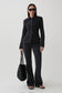A model stands against a light gray studio background wearing a black textured button-front jacket, black wide-leg pants, black sunglasses, and holding a black woven tote bag.