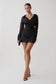 A woman modeling a black long-sleeve wrap mini dress with sheer mesh sleeves and a V-neckline, paired with black high-heeled sandals against a light gray studio background.