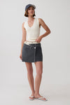 A model stands against a light gray studio background wearing a cream ribbed sleeveless crop top, a dark gray wrap-style denim mini skirt with a white waistband, a navy baseball cap, and white flip-flop sandals.