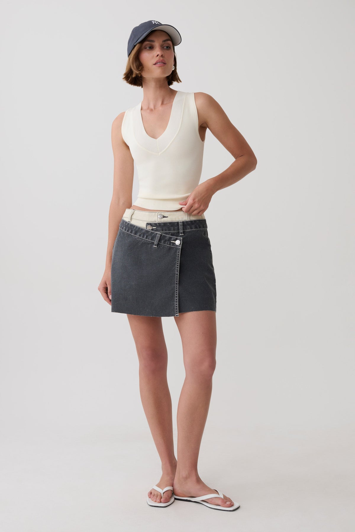 A model stands against a light gray studio background wearing a cream ribbed sleeveless crop top, a dark gray wrap-style denim mini skirt with a white waistband, a navy baseball cap, and white flip-flop sandals.