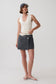 A model stands against a light gray studio background wearing a cream ribbed sleeveless crop top, a dark gray wrap-style denim mini skirt with a white waistband, a navy baseball cap, and white flip-flop sandals.