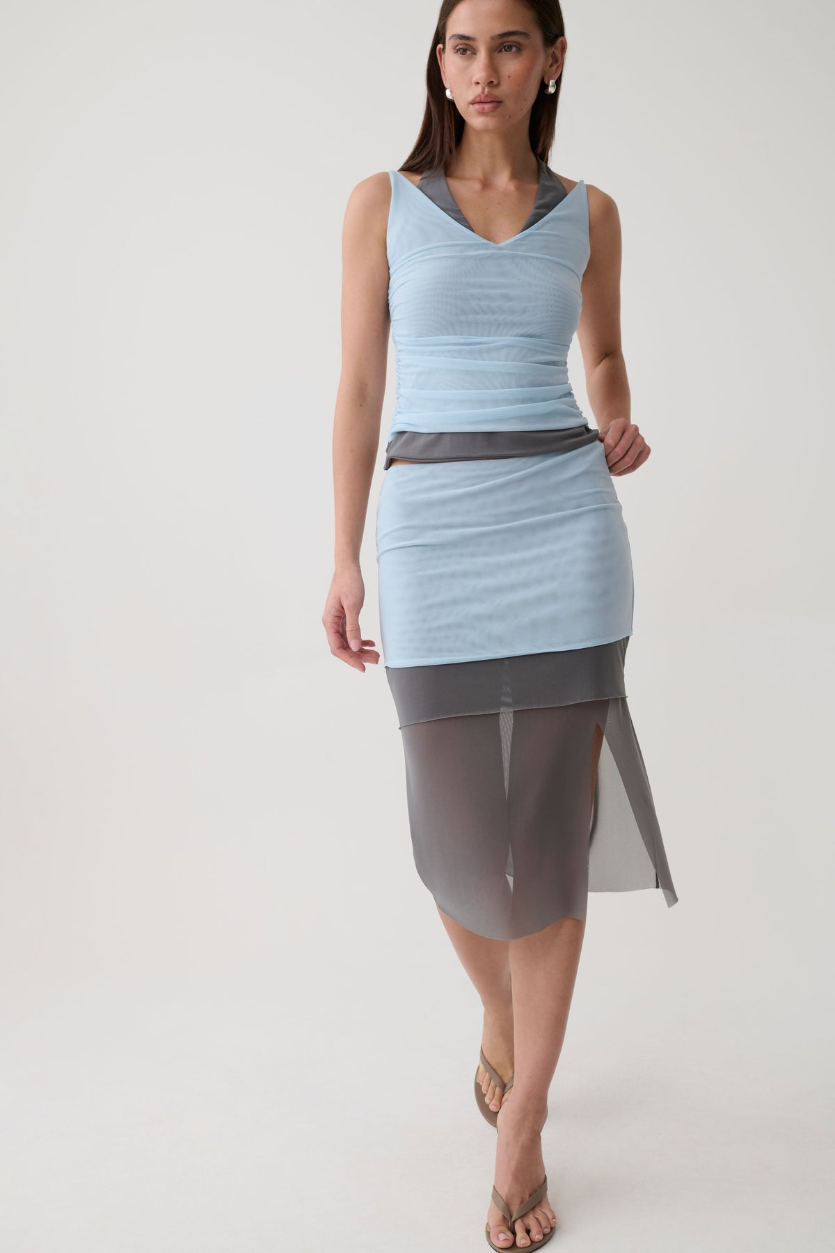 A woman modeling a sleeveless light blue wrap-style dress with a sheer dark gray overlay skirt, photographed against a plain light gray studio background.