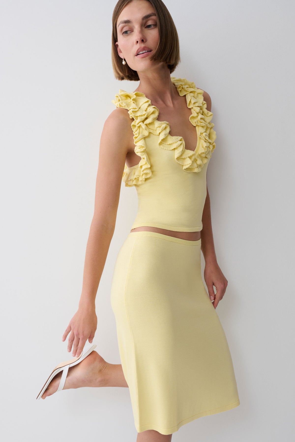 A woman models a pale yellow two-piece knit set consisting of a ruffled halter crop top and a matching midi skirt, paired with white heeled sandals against a plain light gray background.