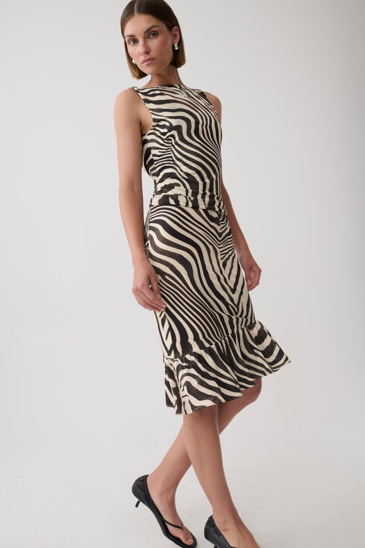A woman modeling a sleeveless halter-neck midi dress with a bold black and beige zebra-like wavy stripe pattern, paired with black heels, against a plain light gray studio background.