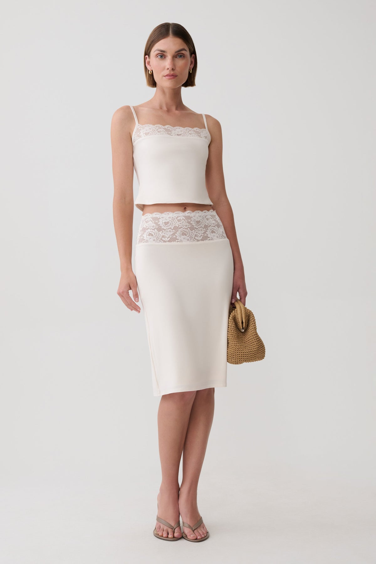 A woman modeling a two-piece cream set consisting of a spaghetti-strap crop top and a matching knee-length skirt with a lace waistband, holding a small woven straw handbag against a light gray studio background.