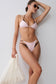 A woman modeling a light pink two-piece bikini with dark brown side mesh details, posing against a light gray wall while holding a white textured beach bag.