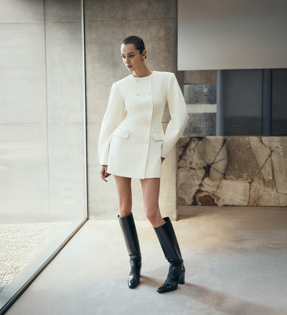 Woman in a white outfit with black boots standing in a modern interior setting.