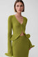 A model wearing a matching olive green knit two-piece set consisting of a cropped cardigan with long sleeves and a high-waisted pencil skirt, shot against a light gray studio background.