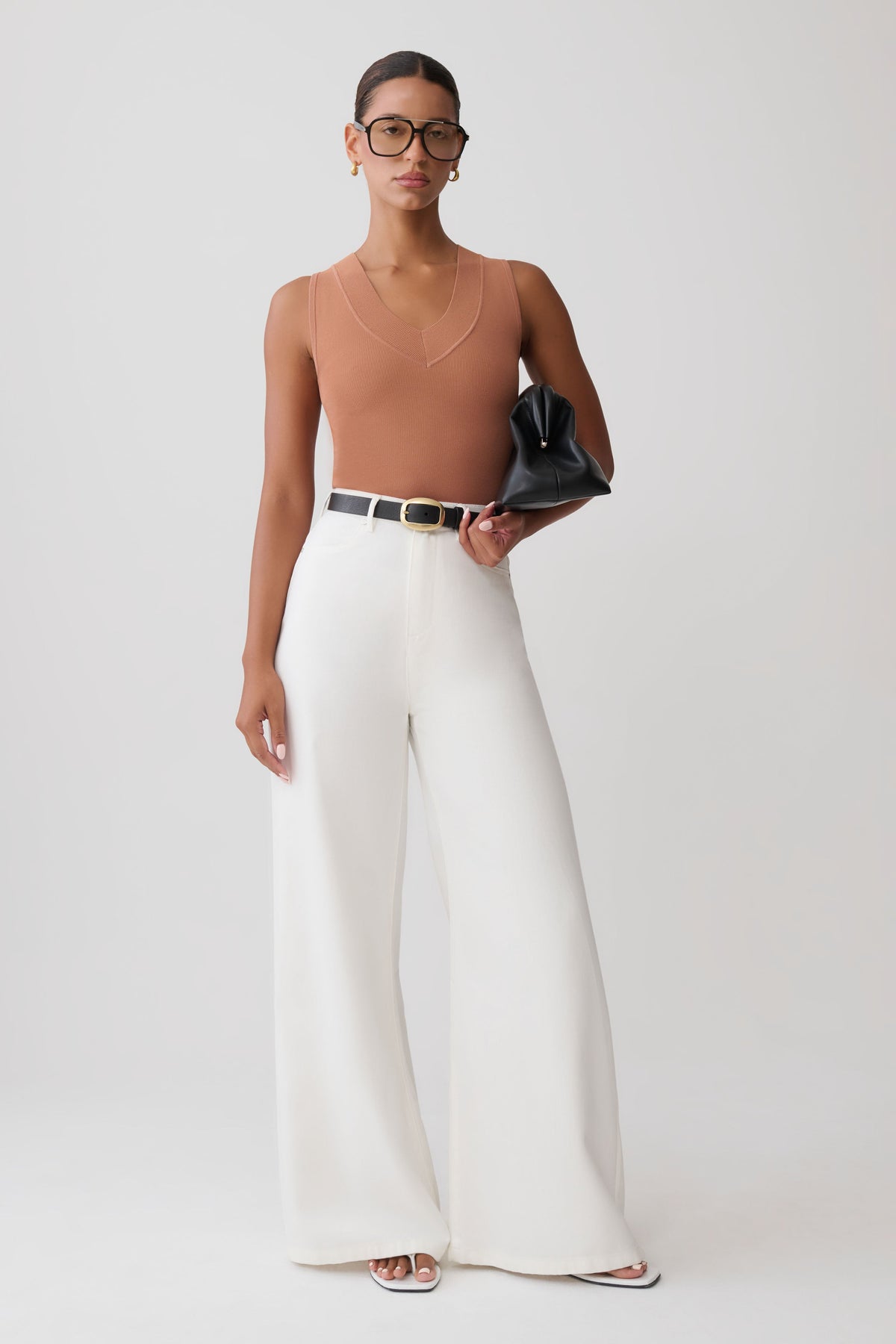 A model stands against a light gray studio background wearing a ribbed tan sleeveless top, high-waisted white wide-leg trousers with a thin black belt, and holding a black clutch.