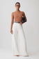 A model stands against a light gray studio background wearing a ribbed tan sleeveless top, high-waisted white wide-leg trousers with a thin black belt, and holding a black clutch.