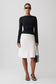 A model stands against a light gray studio background wearing a black long-sleeve top and a white knee-length skirt, holding a small dark gray handbag.