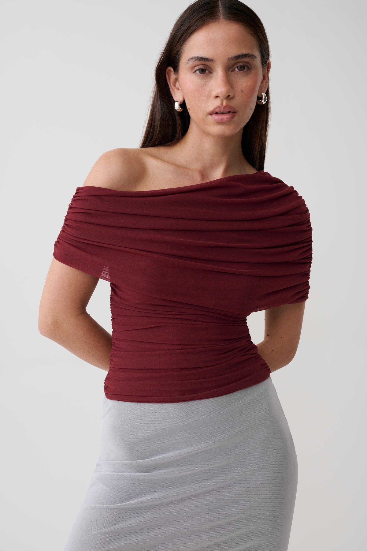 A woman modeling a ruched off-the-shoulder top in deep burgundy, paired with a light gray ribbed skirt, against a plain light gray studio background.