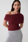 A woman modeling a ruched off-the-shoulder top in deep burgundy, paired with a light gray ribbed skirt, against a plain light gray studio background.