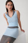 A woman wearing a light blue sleeveless ribbed knit dress with contrasting dark gray trim and a matching gray belt, seated on a white block against a light gray studio background.