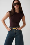 A woman modeling a fitted sleeveless dark brown bodysuit with blue jeans and a gold belt, wearing oversized dark sunglasses against a light gray studio background.