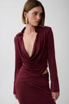 A woman modeling a long-sleeve, deep burgundy wrap-style dress with a plunging V-neckline and gathered draping details, set against a plain light gray studio background.