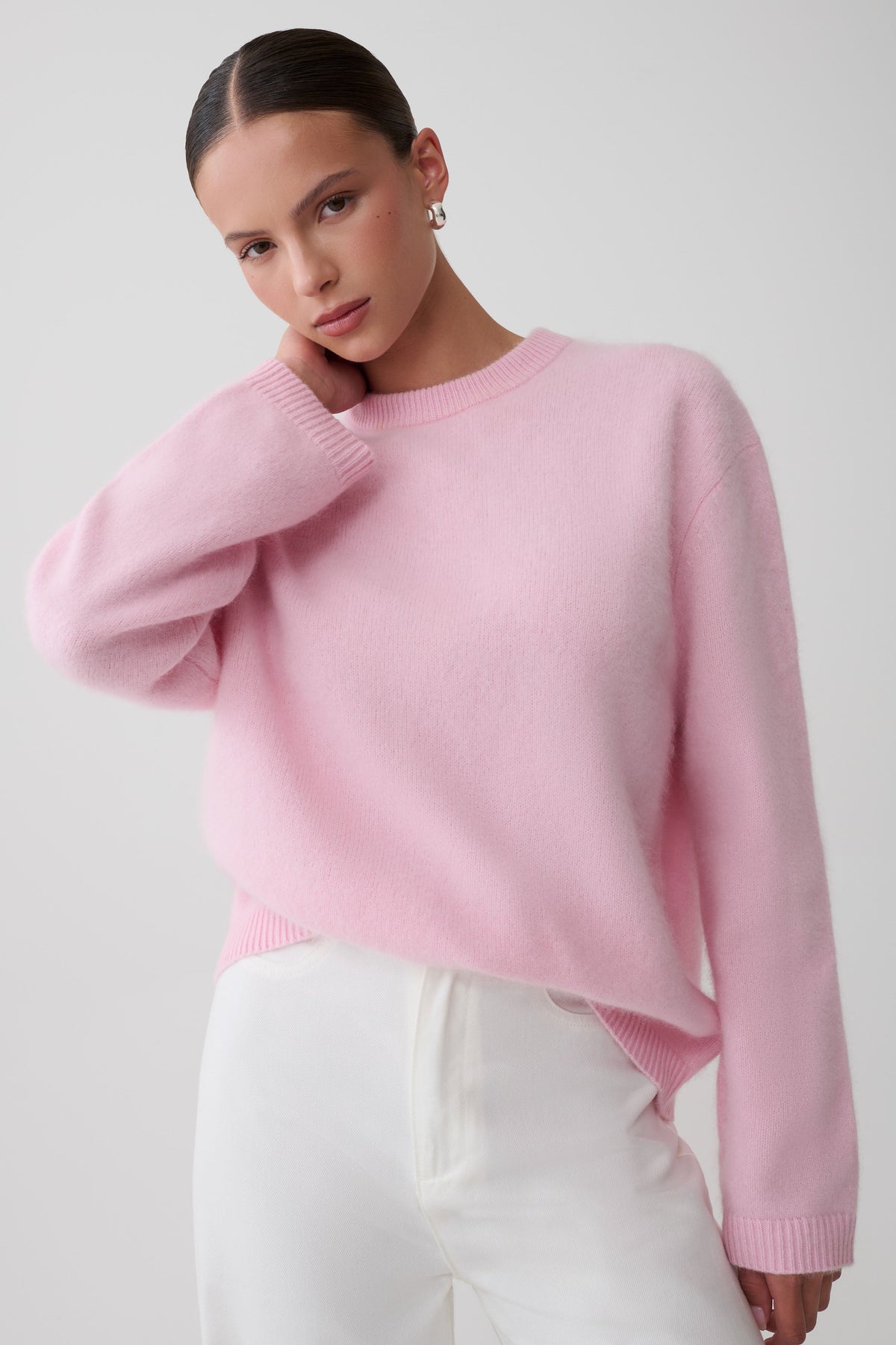 A woman wearing a light pink crewneck knit sweater and white trousers, posing against a light gray studio background.