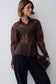 A woman models a sheer, long-sleeved button-up blouse in a dark brown color, paired with dark wide-leg trousers and holding a black textured clutch bag against a plain light gray background.