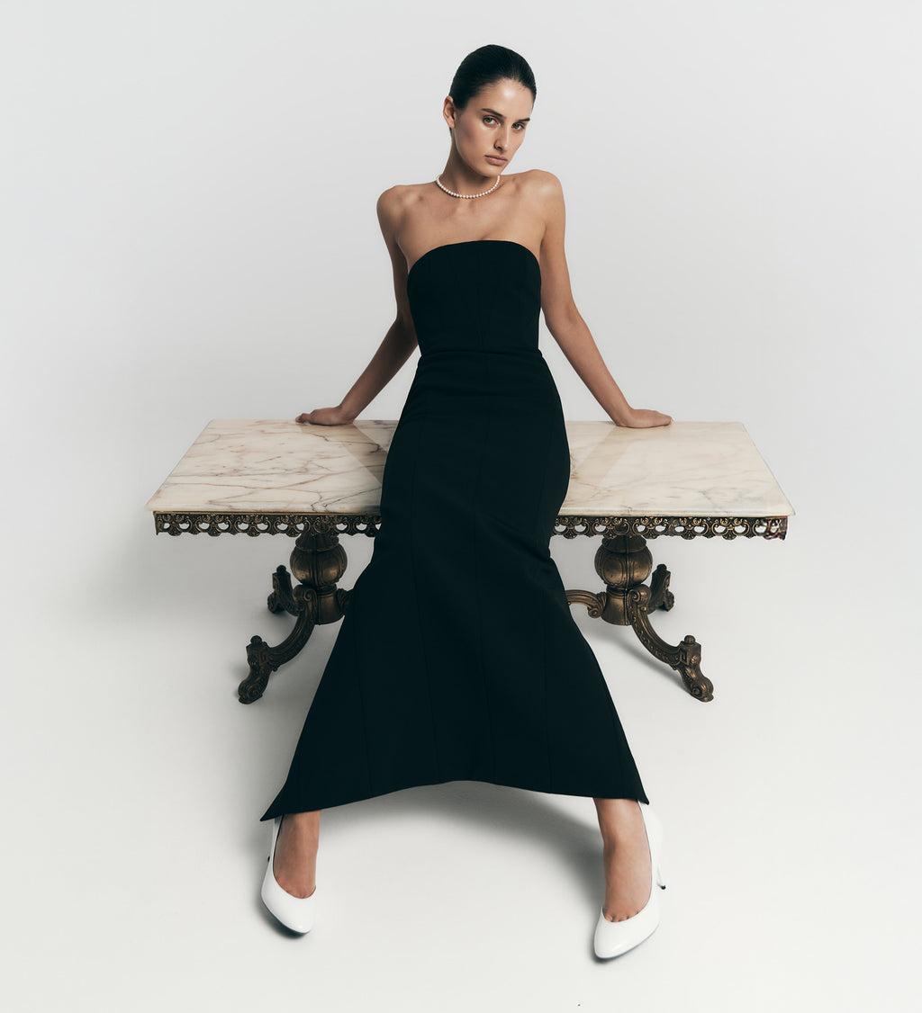 Woman in a black strapless dress sitting on a marble bench against a white background