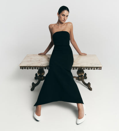 Woman in a black strapless dress sitting on a marble bench against a white background