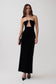 A woman modeling a strapless black velvet maxi dress with a sweetheart neckline and a keyhole cutout at the waist, paired with black pointed-toe heels against a light gray studio background.