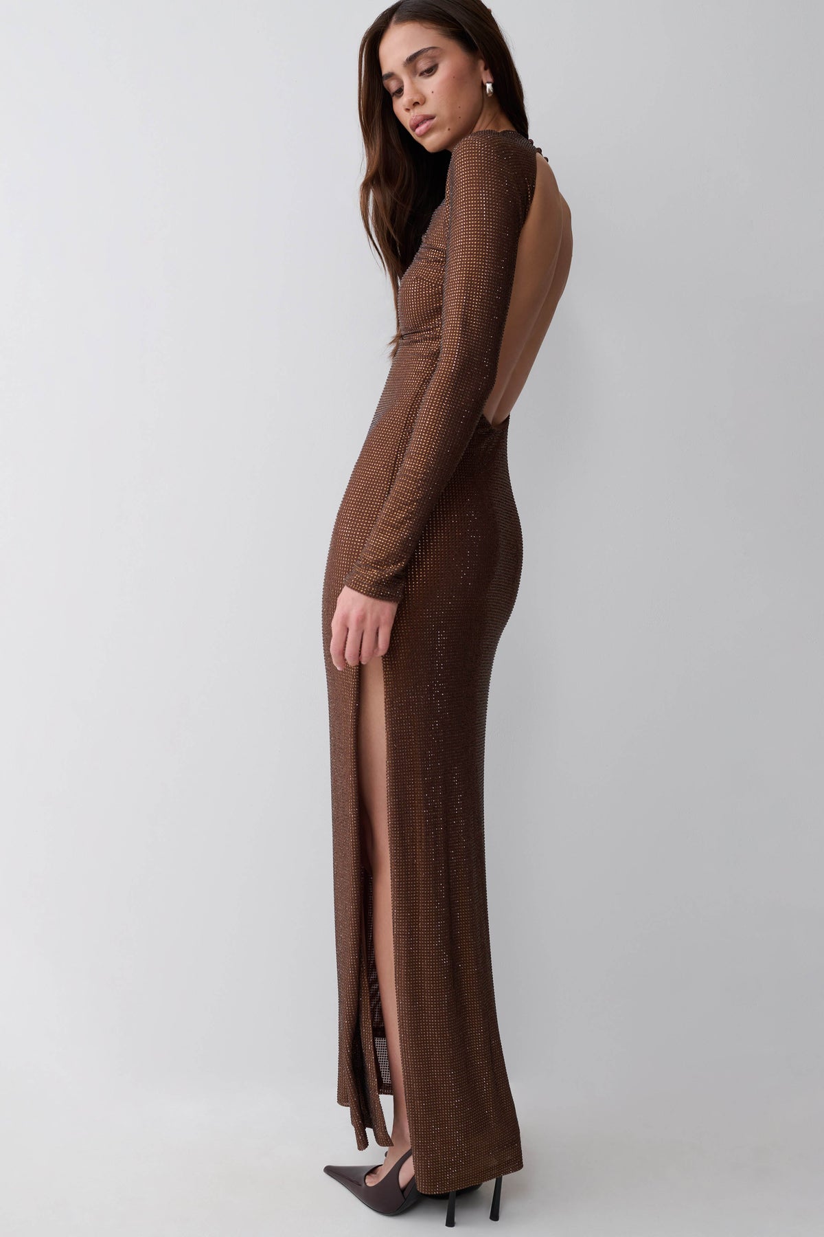 A woman modeling a long-sleeve, floor-length brown knit dress with a high front slit, shown from the back against a light gray studio background.