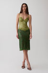 A woman models a green lace midi dress with a corset-style bodice and spaghetti straps, paired with black heeled sandals, against a plain light gray studio background.